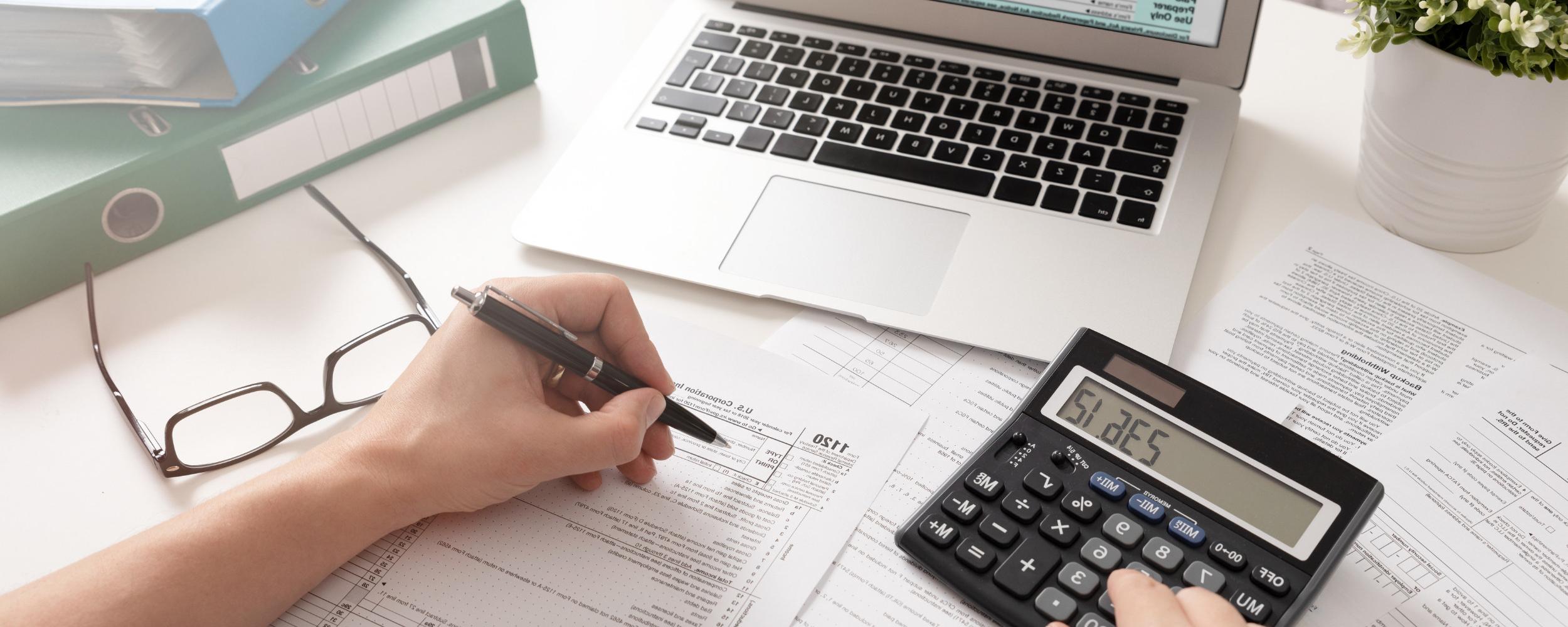 Picture of a calculator, open laptop and the hand of a person taking notes.