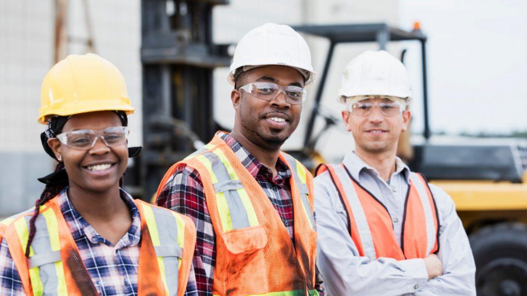 Three people - one white male, one black male, and one black female - all wearing hard hats and orange safety vests stand together smiling.